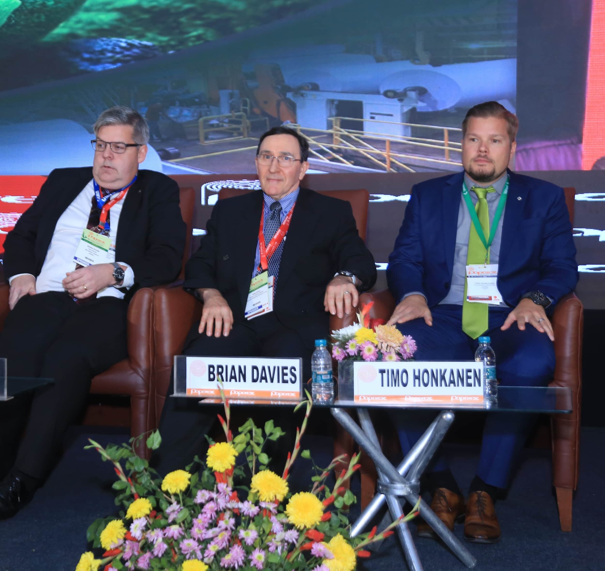 Three men seated at a conference panel with name plates and floral decorations