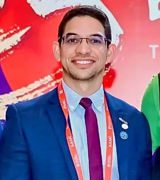 Retrato de Alex Vieira, CIO e VP do HCOR e ABCIS. Um homem jovem de óculos de armação preta, com barba curta e sorriso aberto, vestindo uma camisa social de padronagem xadrez pequena em tons de azul claro e branco.