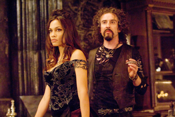 Rosario Dawson as Persephone in a black corset standing by a rock star looking Hades.