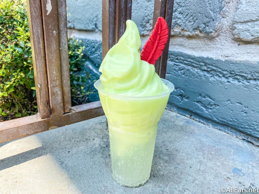 Sitting on a partition in front of the bushes, the light green lime soft serve ice cream sits in a tall cup of sprite, there is a red, chocolate feather sits in the soft serve, resembling Peter Pan's cap.