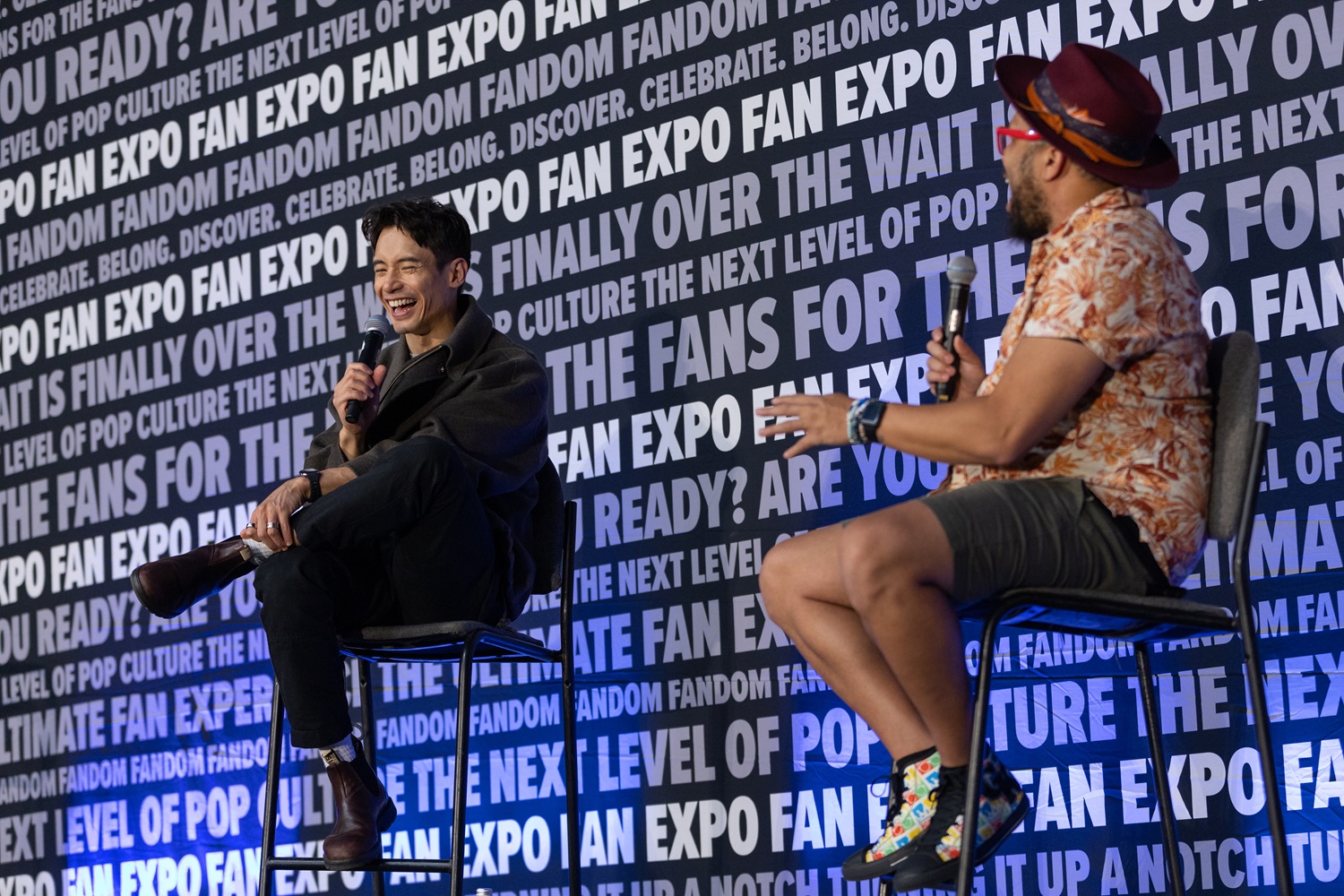 Actor Manny Jacinto in dark clothing with a microphone, laughing during a panel at FAN EXPO. The background shows FAN EXPO branding with various phrases like "Are you ready?" and "The wait is finally over."