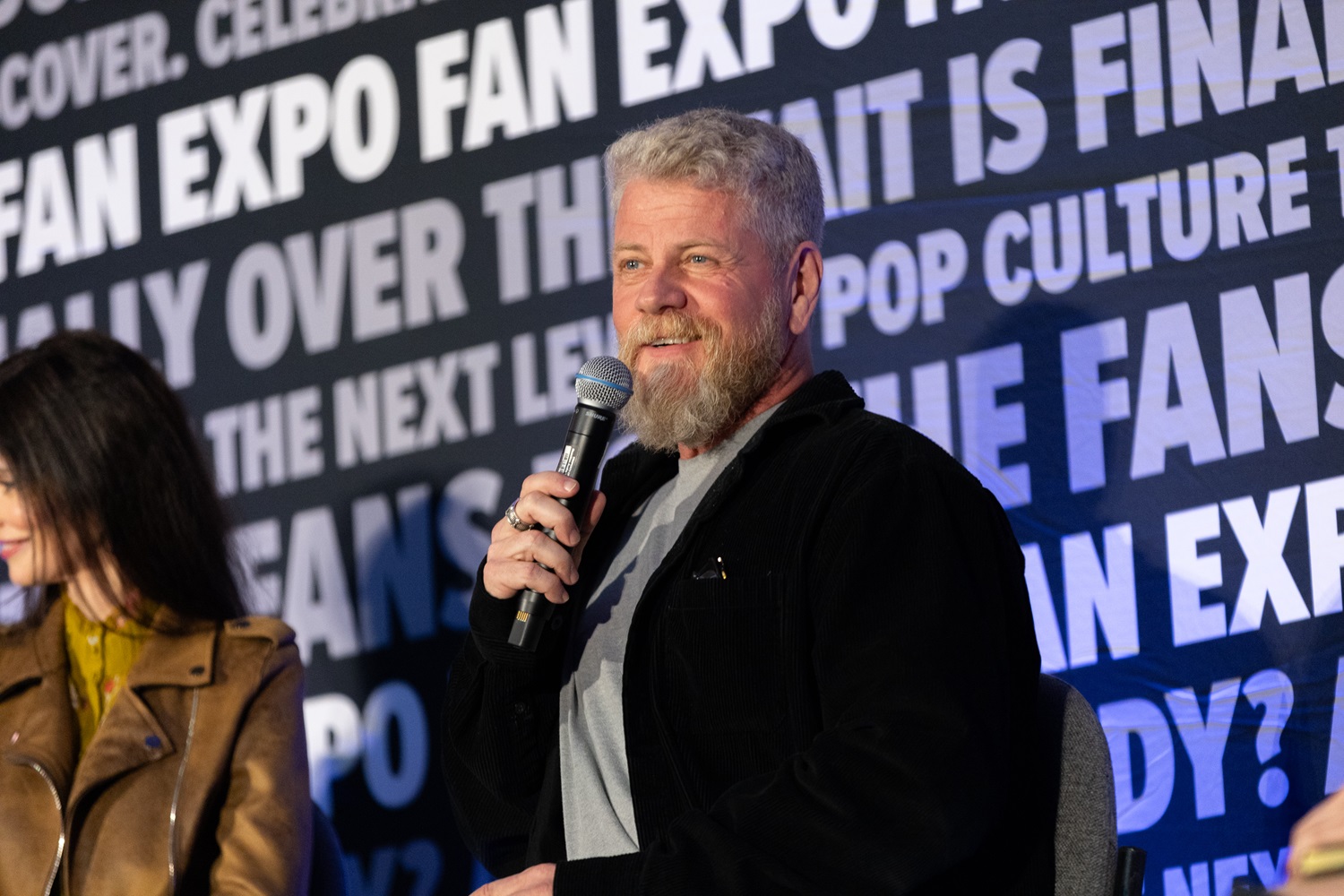 Actor Michael Cudlitz with a light beard, holding a microphone and smiling while speaking at a panel at FAN EXPO. Background features FAN EXPO branding with phrases celebrating fandom and pop culture.