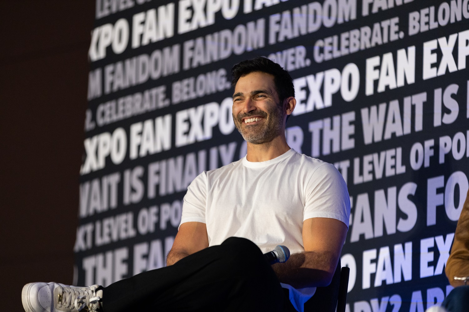 Actor Tyler Hoechlin wearing a white t-shirt, smiling broadly during a panel at FAN EXPO. The background is covered in FAN EXPO branding with enthusiastic phrases about the event.