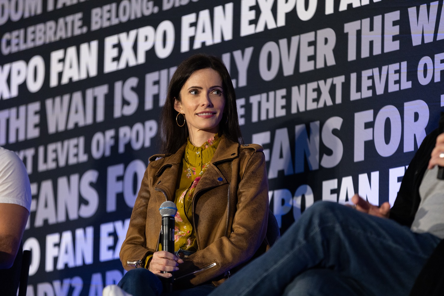 Actress Bitsie Tulloch wearing a brown jacket and floral blouse, attentively holding a microphone while seated on stage at a FAN EXPO panel. The backdrop has FAN EXPO logos and phrases for fan engagement.