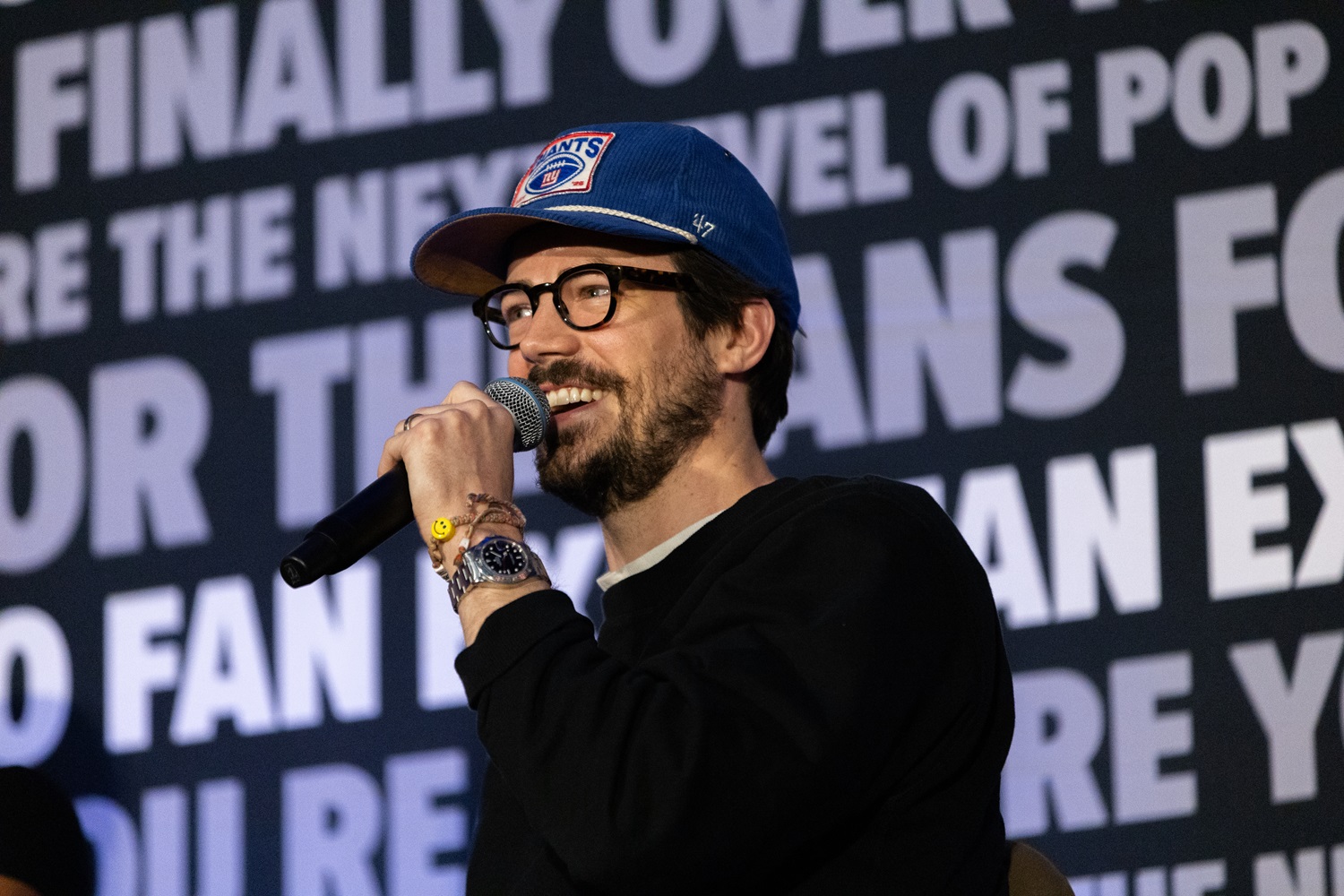 Actor Grant Gustin wearing glasses and a blue cap, holding a microphone and smiling during a FAN EXPO panel. The background is covered with FAN EXPO phrases about pop culture and fandom.