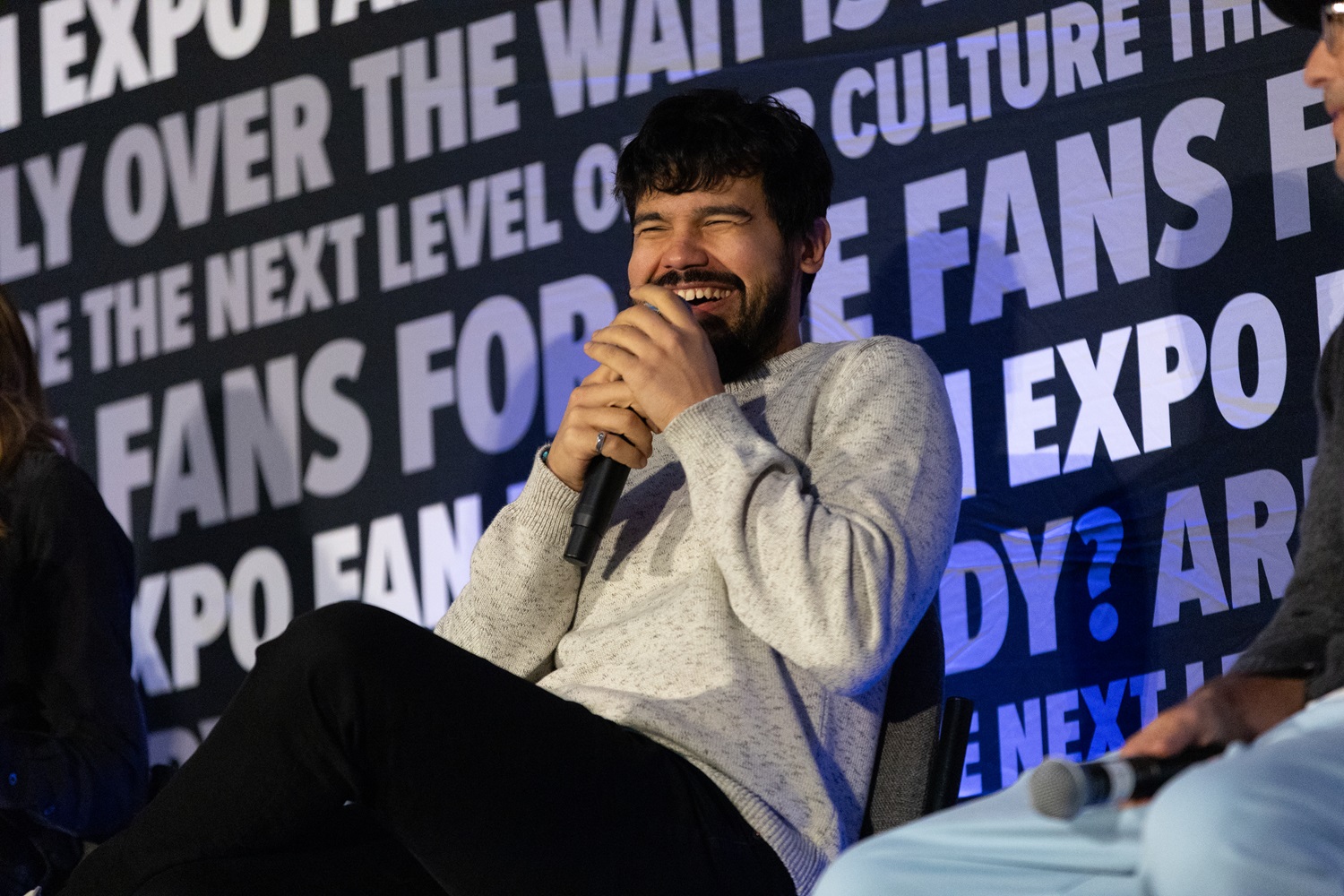 Actor Carlos Valdes with a scruffy beard and sweater, laughing with a microphone in hand during a FAN EXPO panel. The backdrop displays FAN EXPO phrases like "For the fans."