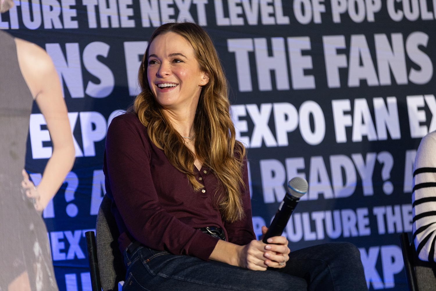 Actress Danielle Panabaker smiling warmly, seated with a microphone on stage at a FAN EXPO panel. The background shows FAN EXPO branding and event slogans.