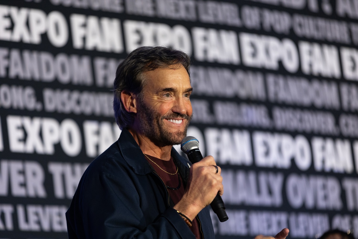 Actor John Wesley Shipp holding a microphone and smiling while speaking during a panel at FAN EXPO. The background has FAN EXPO branding and slogans.