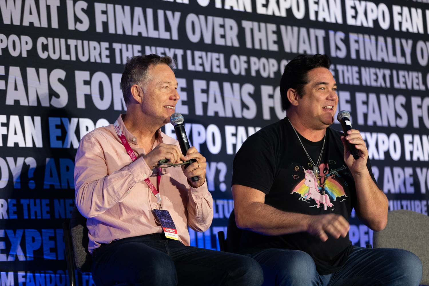 Two actors, George Newbern and Dean Cain, share a lively moment on stage during a panel at FAN EXPO. Both hold microphones. Dean is wearing a t-shirt featuring a unicorn and Deadpool graphic. The background displays FAN EXPO event slogans and logos.