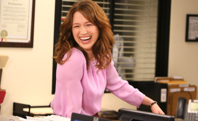 Erin, leaning on another employee's desk, laughs at whatever the person off camera is saying.