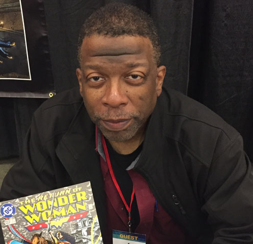 Christopher Priest in a black jacket and red undershirt poses with his Wonder Woman comic