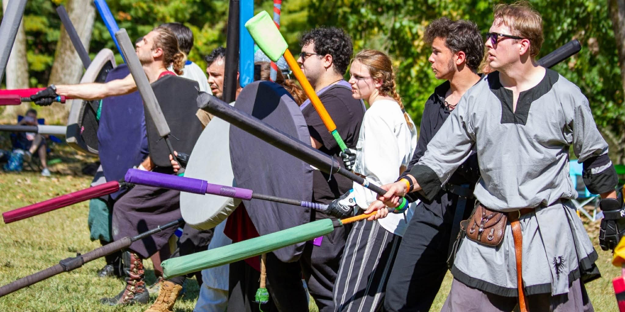 Fantasy reenactors dressed in medieval clothing stand outdoors holding swords and shields. The group prepares for mock combat, representing historical fantasy and medieval role-play at FAN EXPO Cleveland.