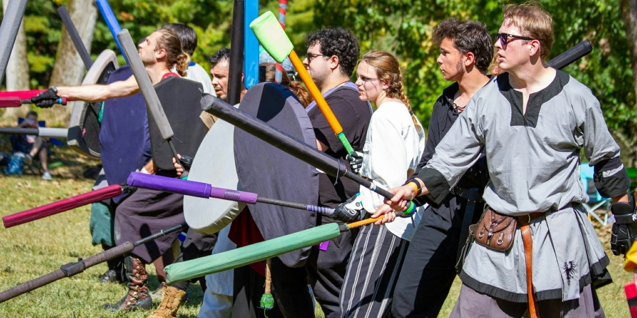 Fantasy reenactors dressed in medieval clothing stand outdoors holding swords and shields. The group prepares for mock combat, representing historical fantasy and medieval role-play at FAN EXPO Cleveland.