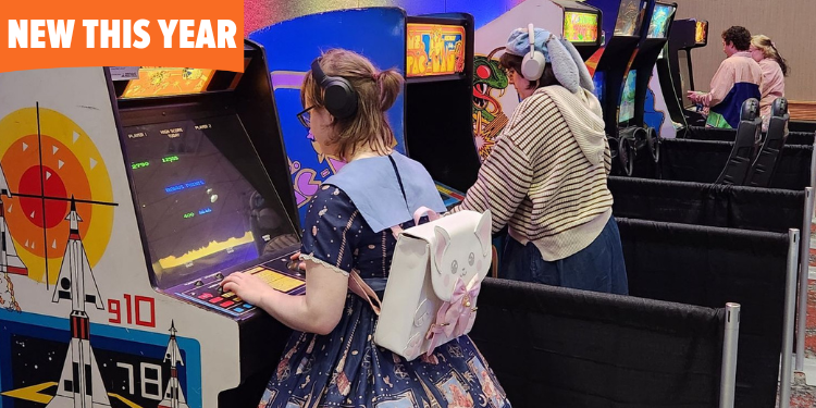 Two fans play vintage arcade games at FAN EXPO Cleveland. One wears a Lolita-style outfit with a pastel backpack, while the other sports a striped hoodie and bunny-eared hat. Classic arcade cabinets line the gaming area.