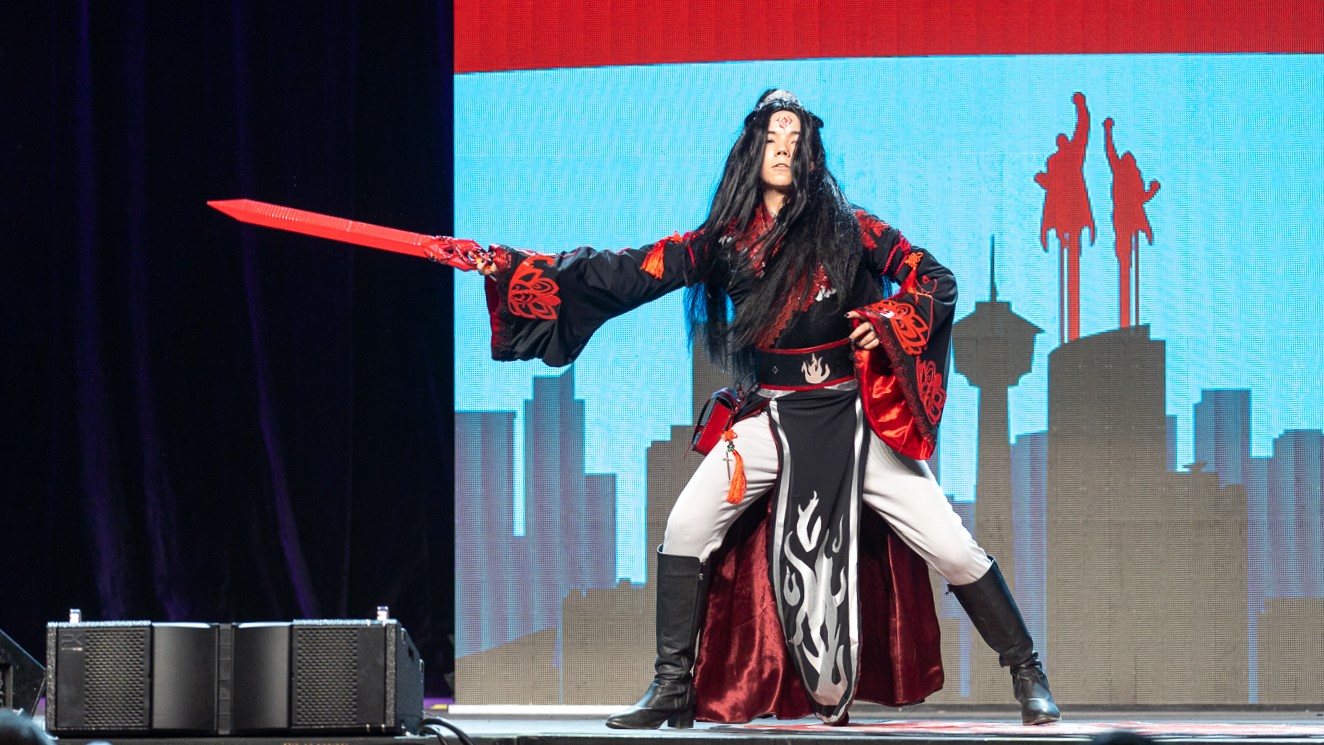 A cosplayer stands on stage holding a red sword. They are wearing a black, red and white costume with flames on it.