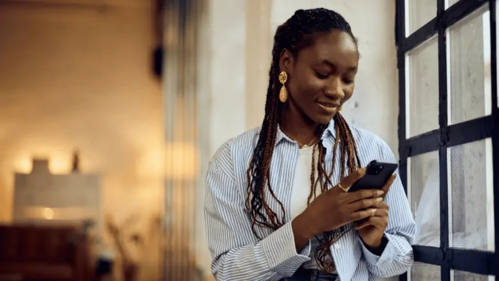 Mulher negra sorrindo com tranças no cabelo, vestindo camisa listrada azul e branca, olha para a tela do celular que segura com as duas mãos enquanto está encostada em uma grande janela de vidro. Imagem sobre o lançamento de concierge digital na Rede Mater Dei.