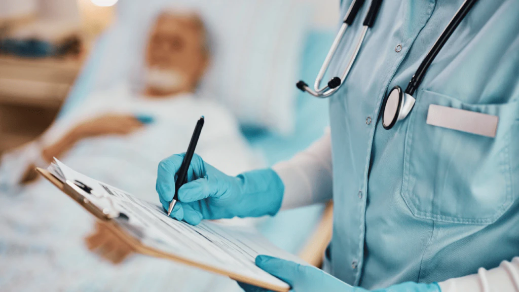 Profissional de saúde com luvas azuis e estetoscópio anota informações em prancheta, com paciente idoso deitado em leito hospitalar ao fundo.