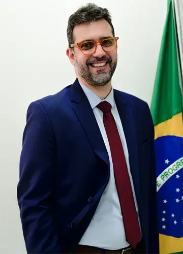 Rodrigo Oliveira sorrindo de óculos de armação alaranjada, terno azul-marinho e gravata vinho, ao lado da bandeira do Brasil.