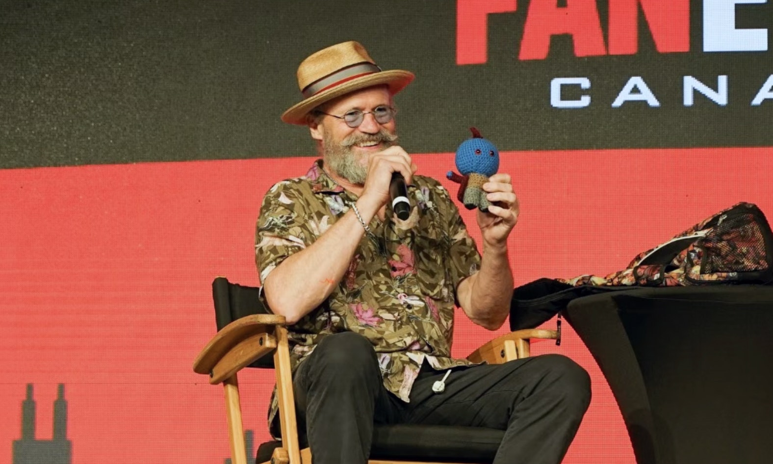 Michael Rooker sits on stage in a hawaiian print blouse with black pants and brown shoes. He is holding up a blue doll during his panel at FAN EXPO Canada.