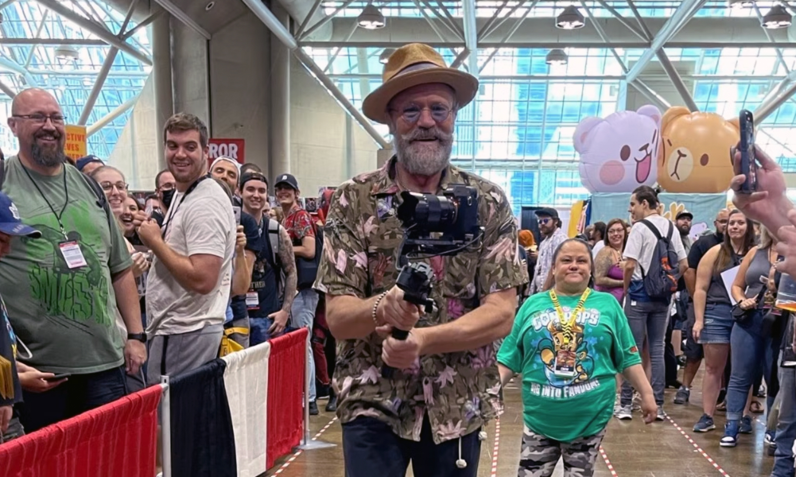 Michael Rooker walks through a crowd of people in a Hawaiian shirt with black pants. He is holding a video camera and smiling.