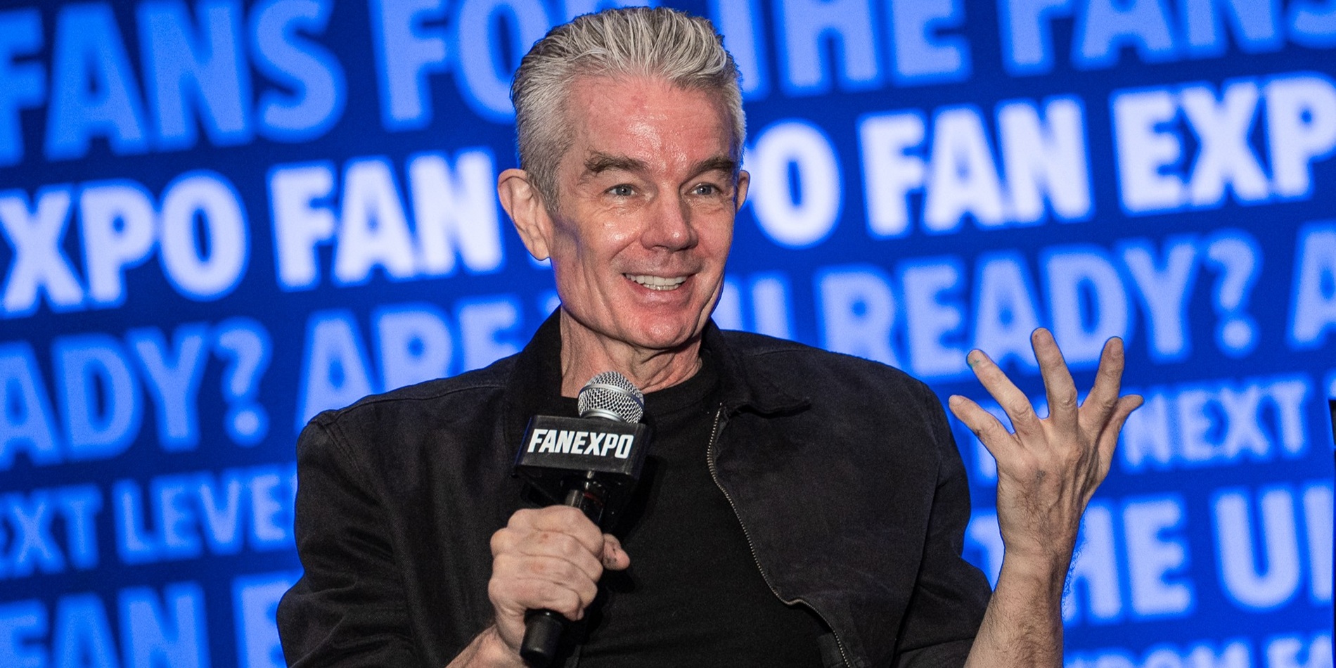 James Marsters speaks on stage at a FAN EXPO Cleveland panel, holding a microphone and gesturing with one hand, with a large blue FAN EXPO-branded screen behind them.