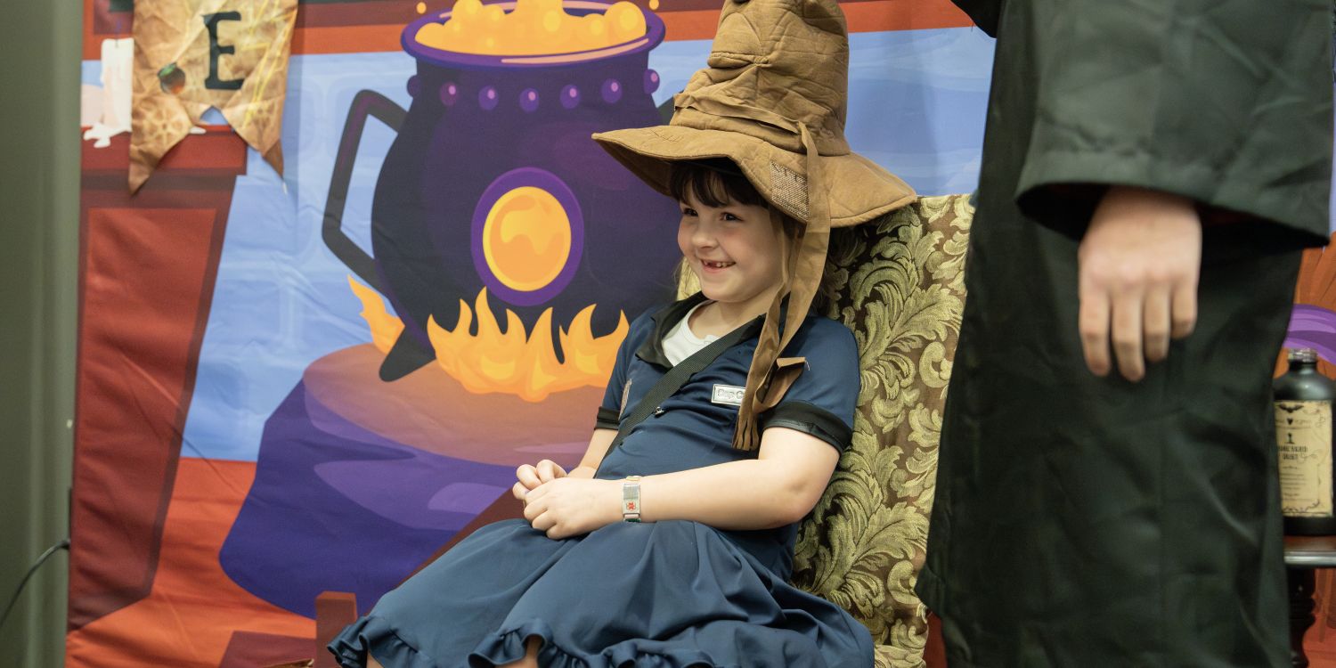 A young girl in a blue dress with a name tag sits on a chair, smiling as she wears a large, wrinkled Sorting Hat from the Harry Potter universe. A colorful backdrop featuring a bubbling cauldron adds to the magical atmosphere.
