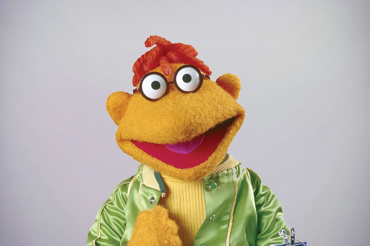 Muppets character Scooter stands in a green jacket, smiling and staring into the camera