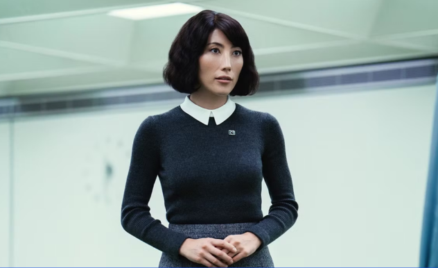 A calm faced woman stands, her hands folded in front of her stomach, and her face indicates she's about to begin speaking to an unseen person.