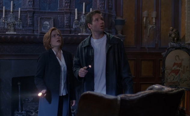 Mulder and Scully hold their flashlights as they look on in awe. They stand in a Victorian style living room.