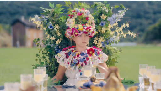 Dani sits at the head of the banquet table, draped in a mass of brightly coloured flowers, contrasted heavily against her white dress. The glasses are filled with a ceremonial wine. Dani's face plays a mix of confusion and concern