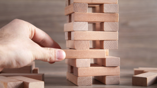 A hand comes in from the left, taking a side piece from the fourth row from the bottom, it is pulled 1/3 of the way out of the stack. There as misc. Jenga pieces strewn around the table