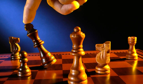 A dimly lit chess board with one of each chess piece in plat. A hand appears from the top of the image, picking up one of the pieces.