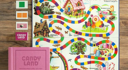 A candy land board laid out with all of the pieces - there are individual cards, a green square, two orange squares, and a chocolate bar, as well as the instruction book
