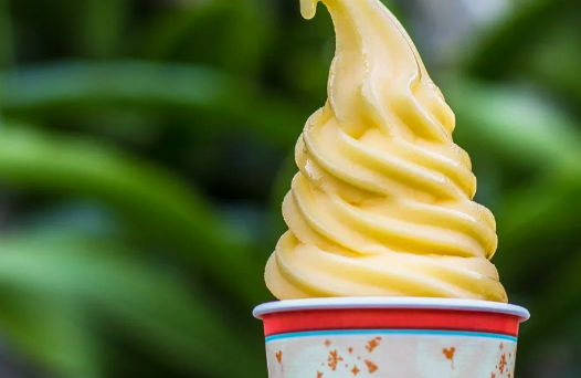 A perfectly swirled, yellow, pineapple dole whip in a disney print cup sits in front of palm leaves