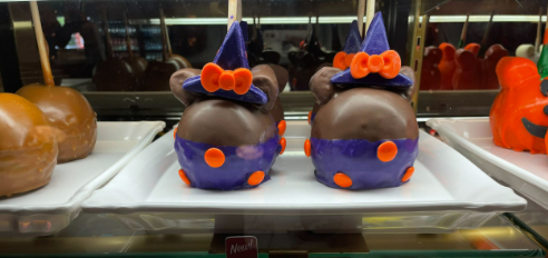 Left, regular caramel apples are plated for purchase. Center, Caramel apples dipped to look like a Witch Minnie Mouse, in a purple dress with orange polka dots. Adhered with more chocolate, a chocolate witches hat with a bow sits on top. Right, Apples dipped in orange candy to resemble Mickey pumpkins, like the statue in the parks.