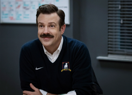 Ted smiles a closed lip smile as he leans over his desk