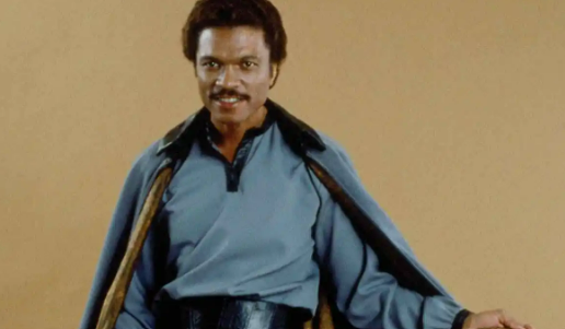 Lando stands in front of an all beige backdrop in his signature blue suit.