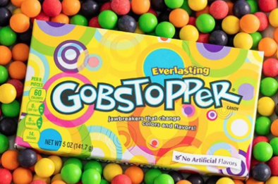 A box of Gobstoppers (yellow, with purple, pink, blue, and white circles of varying colour formations) lays on top of a pile of Gobstoppers. The candies are purple, yellow, orange, green, or red.