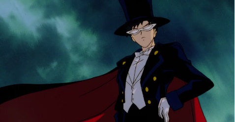 Tuxedo Mask stands, with this hand on his hip and cape blowing in the wind, with the sky behind him.