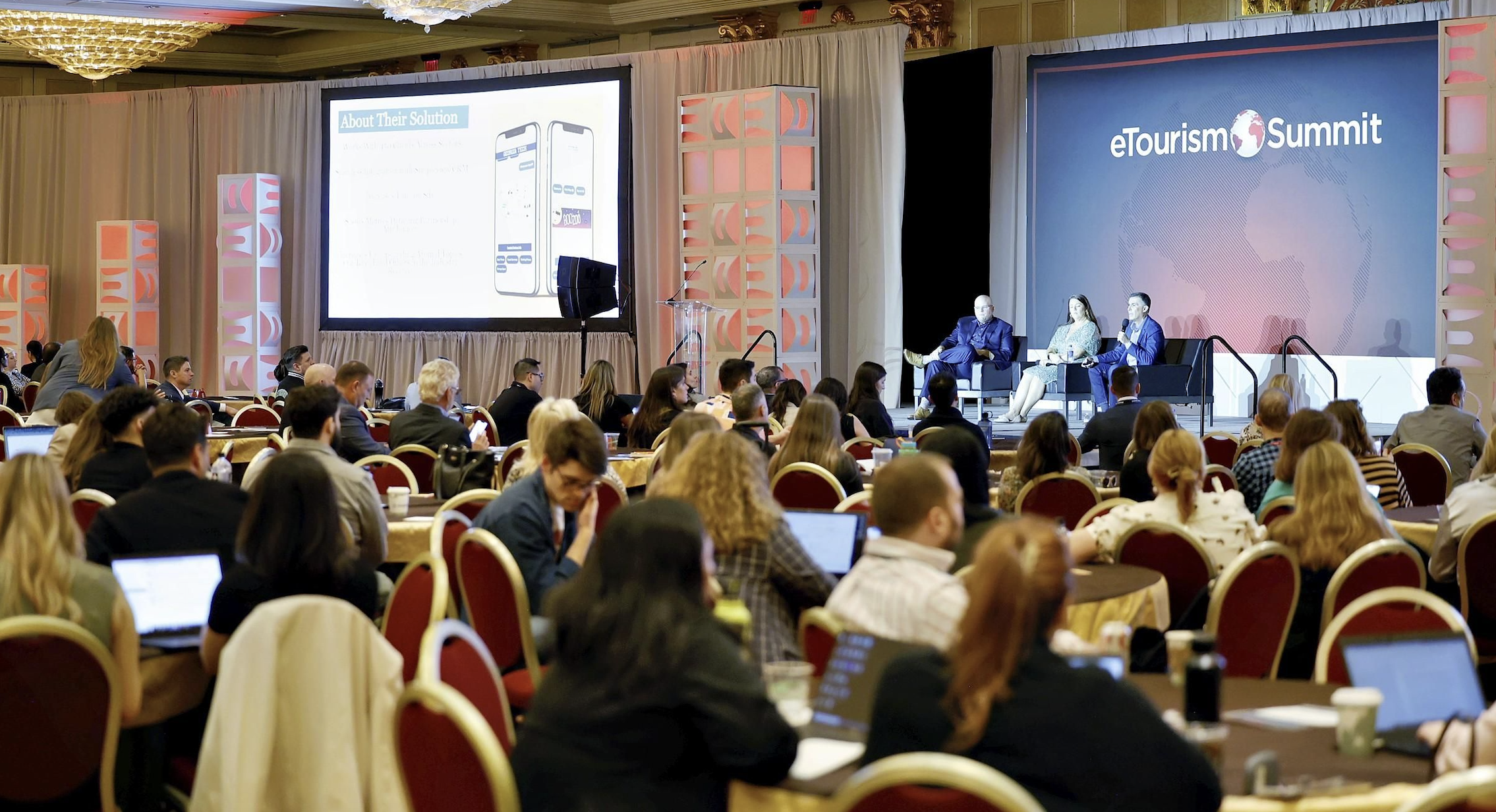 A large ballroom with eTourism Summit attendees seated around tables and three presenters on stage leading an education session.
