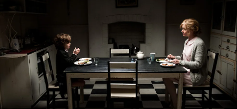 Amelia and Samuel sit across from each other at the dinner table. All of the colour in the room is muted, including the black and white checkered floor, and the black and white table and chairs.