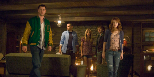 A group of young people stand in the living room of the cabin, staring in horror at something off screen.