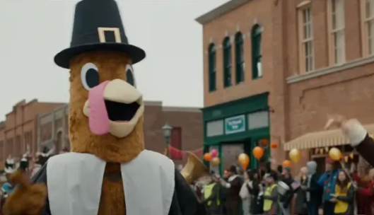 A crowd cheers at the thanksgiving parade. In the foreground, a turkey mascot marches.