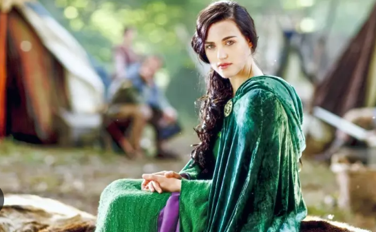 Wearing all green, Morgana sits on a rock amongst tents.