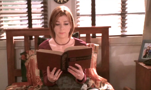 Holding a large book, Willow reads on her bed - she studies the pages with a stern expression.