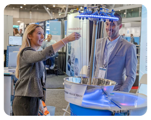 A woman and a man engage in conversation at a bustling trade show, surrounded by various exhibition booths