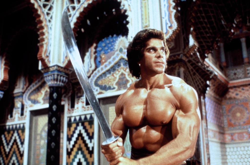 In a room with decorative tiles and arches, Lou is shirtless and is holding a sword, looking off to the right.