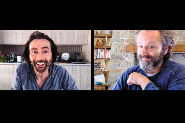 Screen grab of a zoom like call between David Tennant on the left and Michael Sheen on the right. David looks to be in a kitchen area with the sink and a bunch of appliances. Michael Sheen is in a living room area with a stone firepit and a book shelf full of books, pictures and toys.