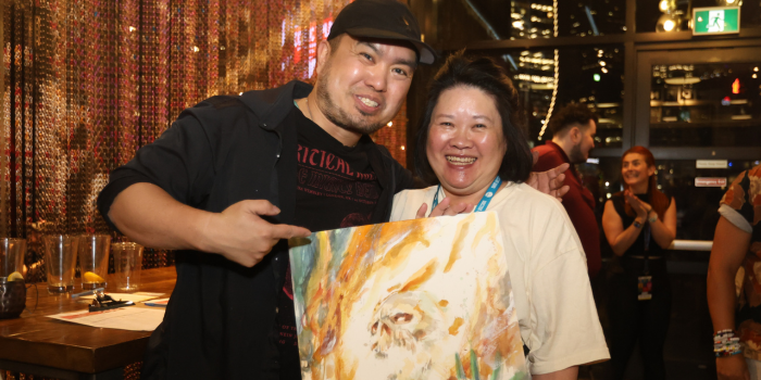 Tony Moy poses with the auction winner of his hand painted Ghost Rider piece at the Rec Room in Toronto. Both smile, the fan, right, smiles as they hold the painting, and Tony, left, smiles and points to the painting.