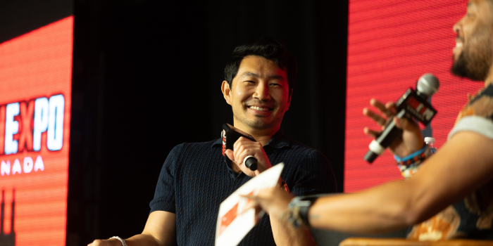 Simu Liu smiles as moderator Victor Dandridge speaks, asking his next question. Both are holding FAN EXPO Canada microphones, and sitting in front of a red and black FAN EXPO Canada background on LED screens.